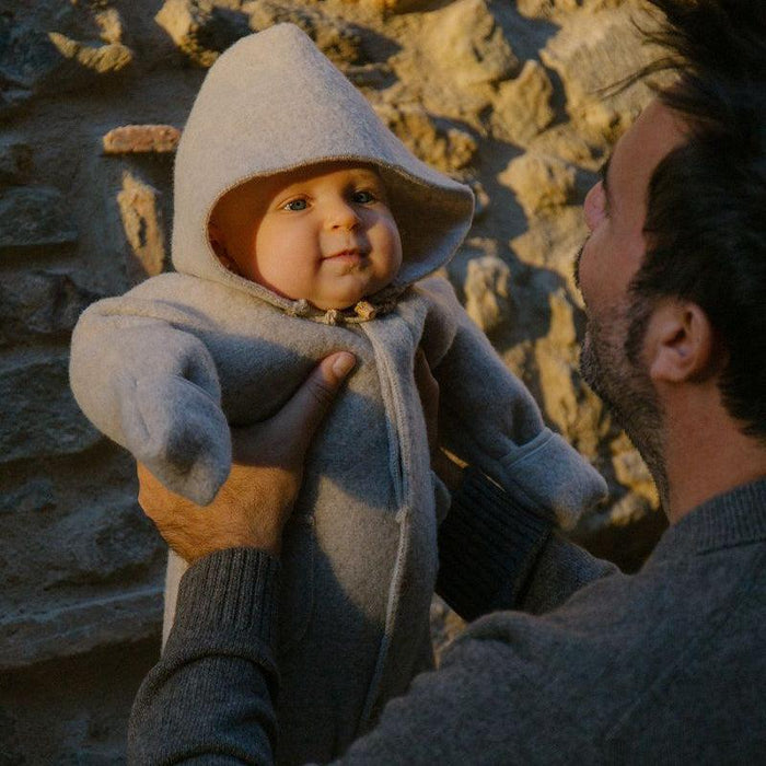 A baby wearing a light gray hooded knit romper is held up by an adult outdoors against a stone wall, with warm sunlight casting shadows.
