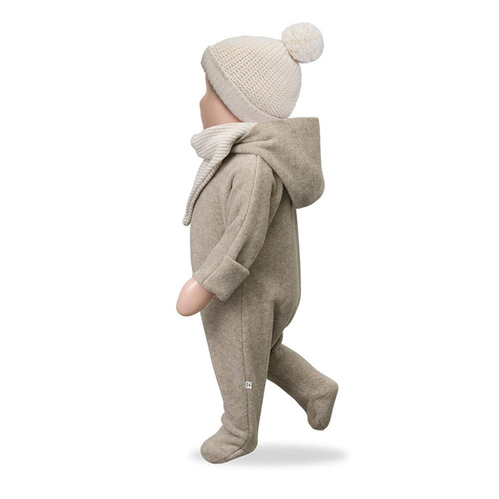 A baby doll or mannequin wearing a beige footed one-piece outfit with a hood, a matching knit beanie with a pom-pom, and a ribbed knit scarf, shown from the side against a white background.