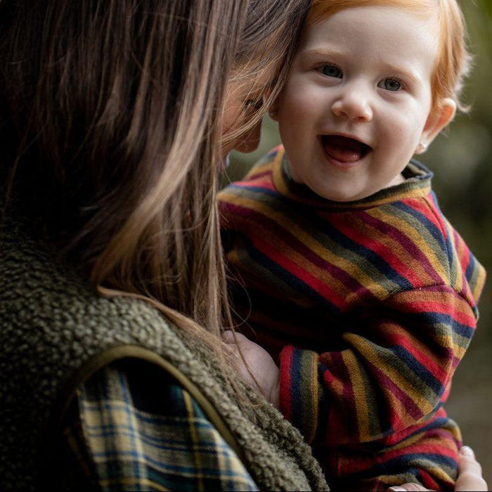 A close-up lifestyle shot of a smiling toddler with red hair being kissed on the cheek by a woman outdoors. The toddler is wearing a colorful striped knit sweater in shades of red, yellow, blue, and green.