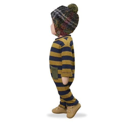 A toddler shown from the side wearing a cozy knit one-piece jumpsuit with mustard yellow and navy blue horizontal stripes, a matching knit pom-pom beanie, and tan shoes, photographed on a clean white background.