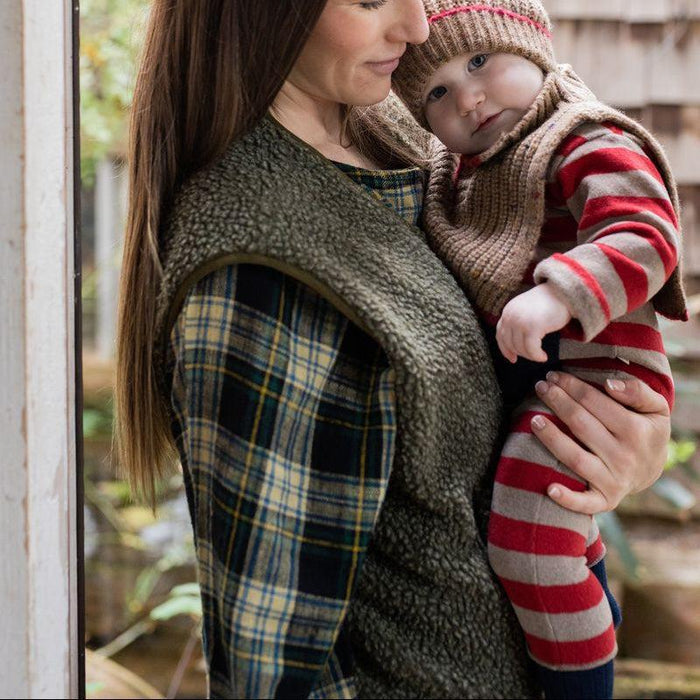 A woman wearing a dark olive green fleece carrier vest holds a baby dressed in a red, beige, and gray striped outfit and a knit bonnet, set against a softly blurred rustic outdoor background.