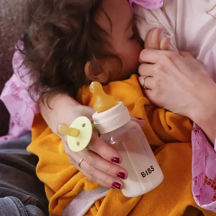 Baby holding a BIBS bottle and pacifier with a person's hand supporting the baby.