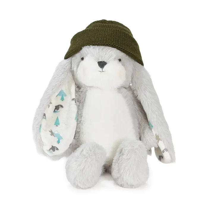 A plush white bunny toy with long floppy ears, wearing an olive green knit hat. The left ear has a printed patch with small blue trees and gray animals, and a small fabric tag is visible on the right ear.
