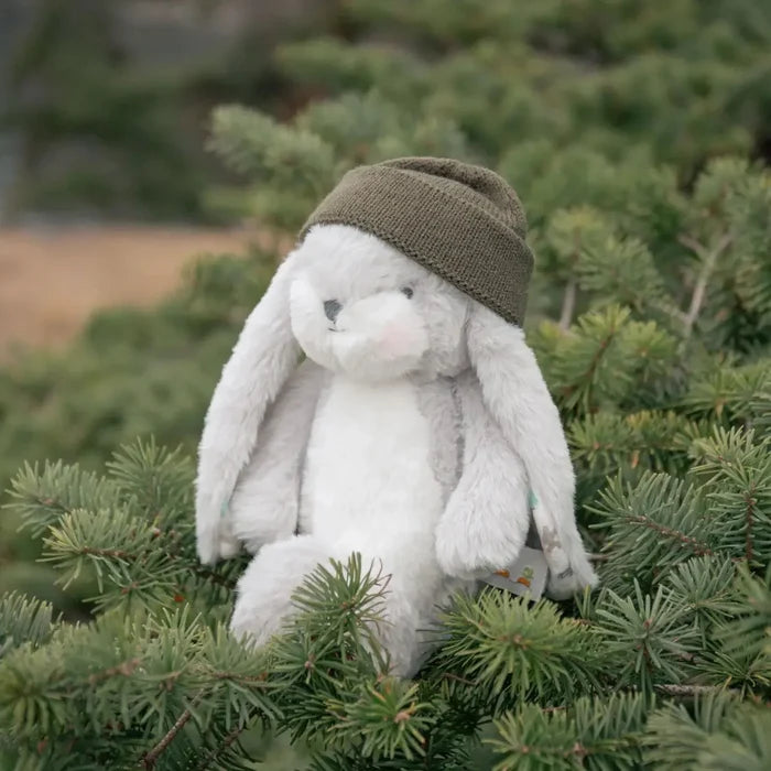 A plush white bunny toy wearing an olive green knit beanie, sitting among green pine branches in an outdoor setting.