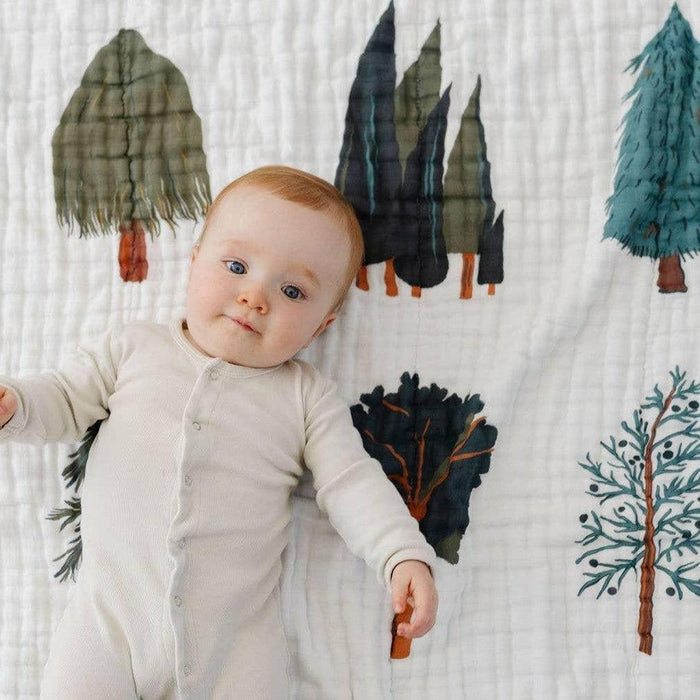 A baby lying on a white muslin blanket printed with illustrated trees in muted green, blue, and brown tones. The baby is wearing a light cream footed one-piece sleeper.