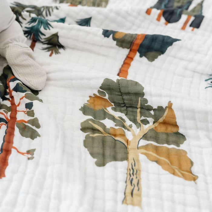 A close-up of a white muslin blanket featuring a printed pattern of stylized trees in muted green, brown, and orange tones, with a white textured glove holding the corner.