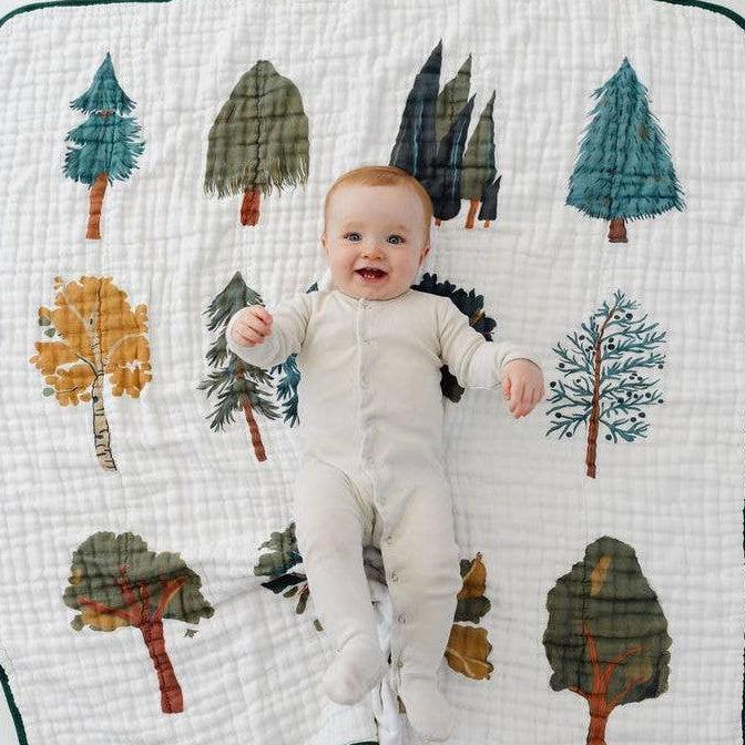 A baby lying on a white muslin blanket with a forest tree pattern, wearing a light cream footed sleeper. The blanket features various stylized trees in green, blue, and orange-brown tones with a dark green border, set against a plain white background.