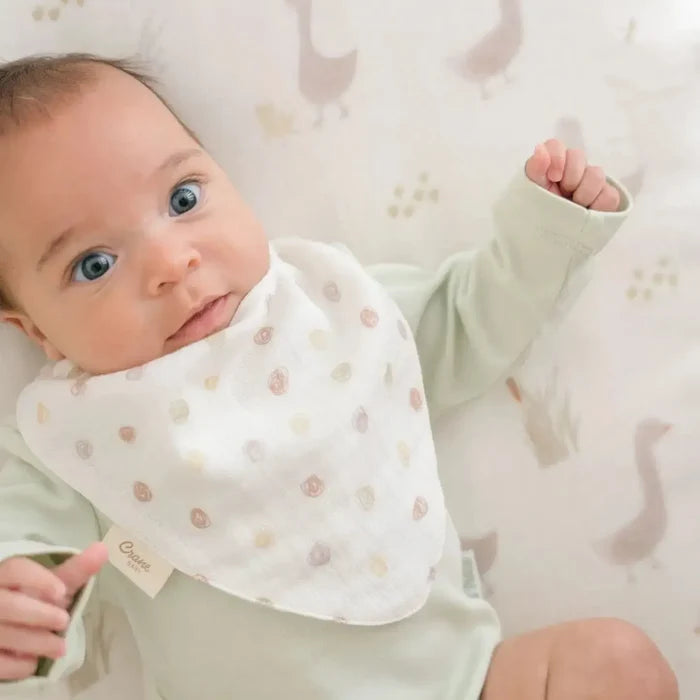 A close-up of a baby wearing a white muslin bib with a small pastel pattern, lying on a patterned changing mat.