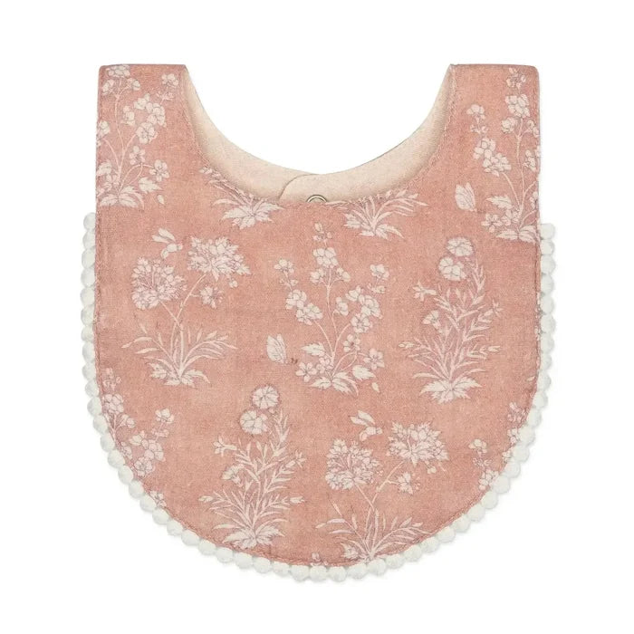 A dusty pink velvet baby bib with a white floral toile pattern and white pom-pom trim along the edges.