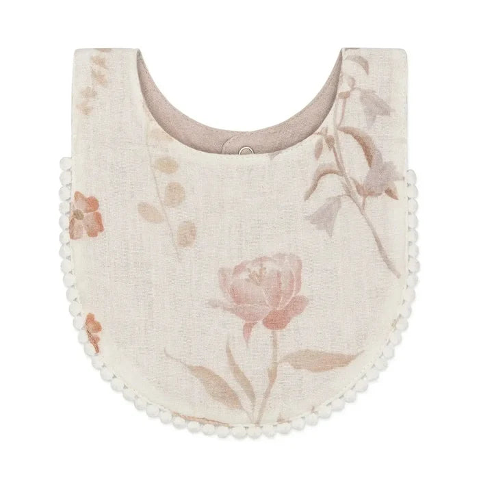 A cream-colored fabric baby bib with a dusty pink floral print and a white pom-pom trim along the curved edge, shown on a plain white background.