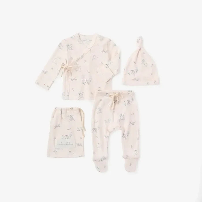 A baby clothing set laid out on a white background, including a long-sleeve wrap top, pants, a beanie, and a small pouch. The set is pale blush pink with a delicate light blue bunny print.