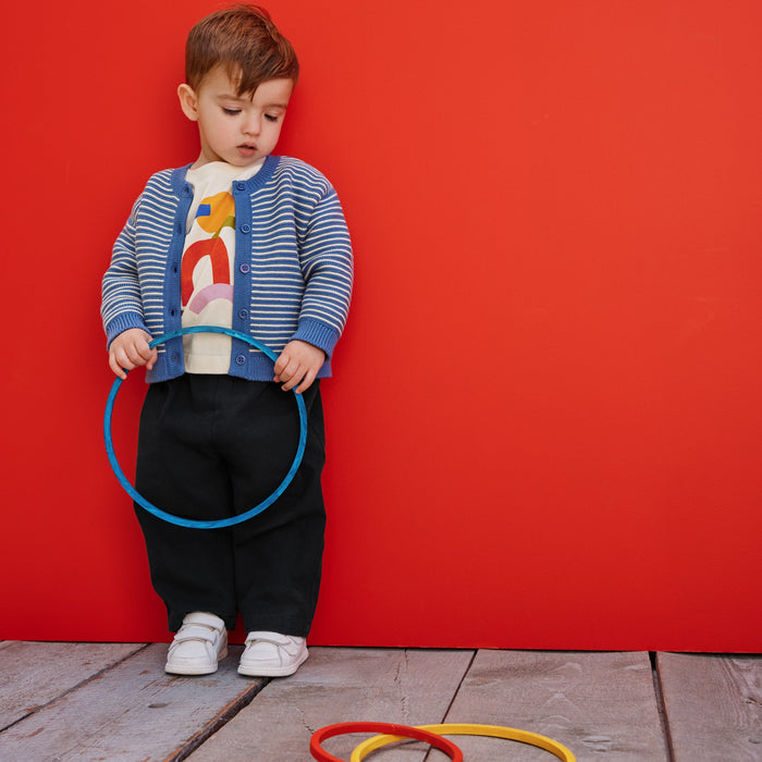 A toddler stands against a bright red wall on a wooden floor, holding a blue hula hoop while two more hoops lie on the floor in front. The child wears a blue-and-white striped cardigan over a white shirt with a colorful graphic, black pants, and white shoes.