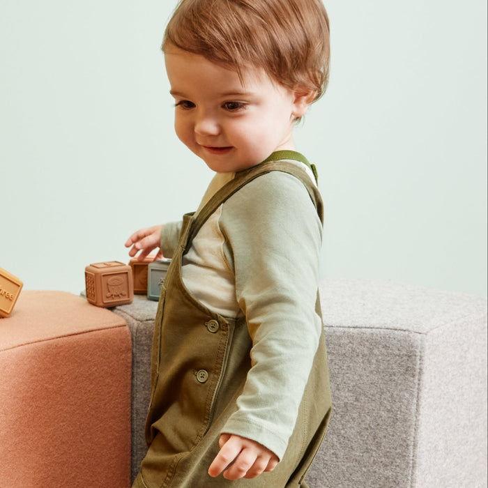 A toddler wearing sage green and white striped overalls walks beside a dusty pink cube ottoman in a minimalist room with a light green wall and wooden floor.