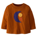 A toddler's long-sleeve crewneck t-shirt in a warm rust-orange color, featuring a cute graphic of a smiling dark blue crescent moon, a yellow sun, and a red star on the front.