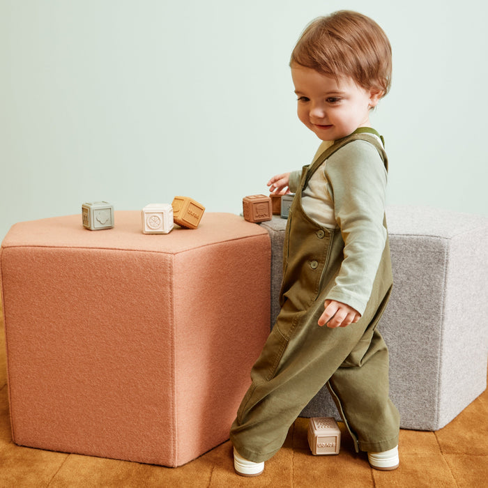 A toddler wearing sage green and white striped overalls walks beside a peach-colored cube ottoman in a minimalist room with a light green wall and wooden floor.