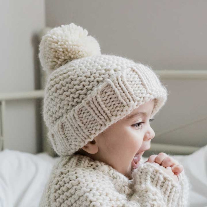 Child wearing a knitted beige hat with a pom-pom, sitting on a bed.
