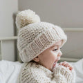 Child wearing a knitted beige hat with a pom-pom, sitting on a bed.