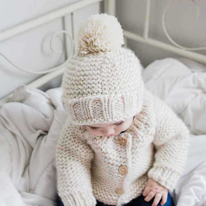 Child wearing a cream-colored knitted hat and cardigan sitting on a white bed.