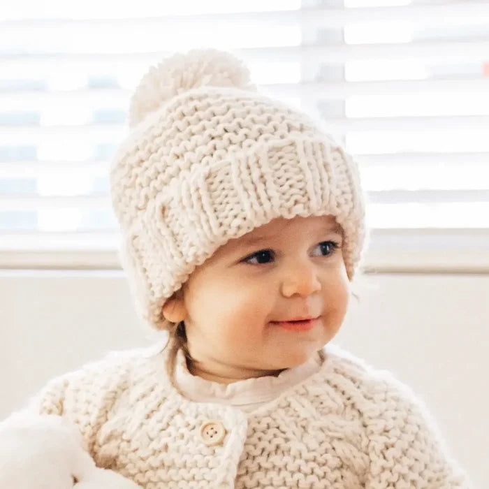 Baby wearing a cream-colored knit hat with a pom-pom, sitting indoors.