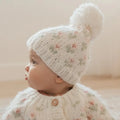 A baby wearing a cream-colored knit hat with a matching pom-pom and a matching cream knit cardigan, both decorated with a delicate pink and green floral pattern. The baby is shown in profile against a soft, warm beige background.