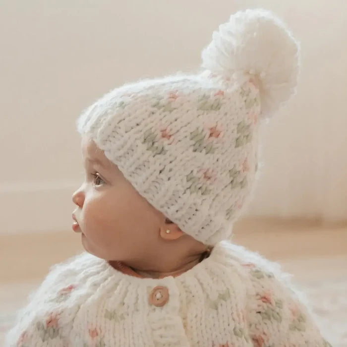 A baby wearing a cream-colored knit hat with a matching pom-pom and a matching cream knit cardigan, both decorated with a delicate pink and green floral pattern. The baby is shown in profile against a soft, warm beige background.