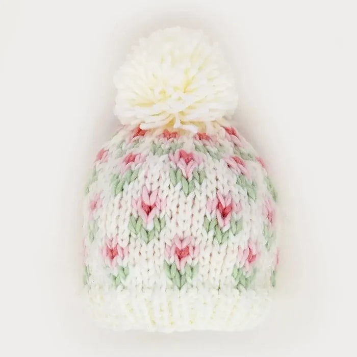 A cream-colored knit beanie hat with a fluffy cream pom-pom on top. The knit pattern features small pink and green tulip motifs throughout the crown.