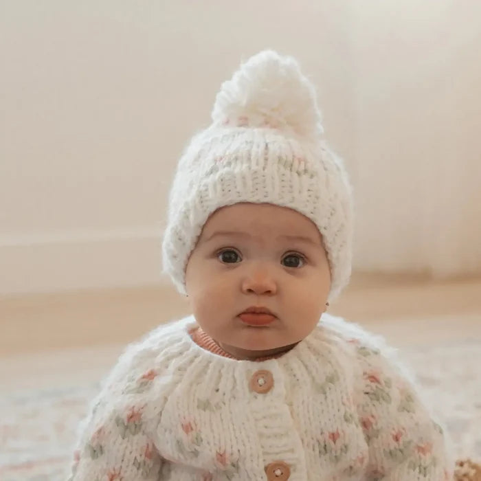 Baby wearing a white knitted hat and sweater with floral pattern on a neutral background