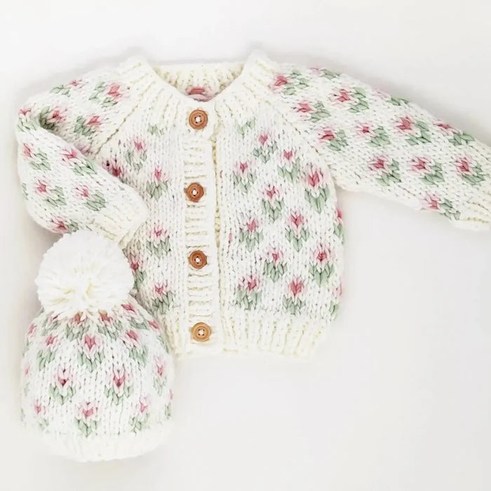 A cream knit baby cardigan and matching hat set with a pink and green floral pattern, displayed on a white background with a black banner at the top reading 'search ’bitty blooms’ to find the coordinating sweater'.