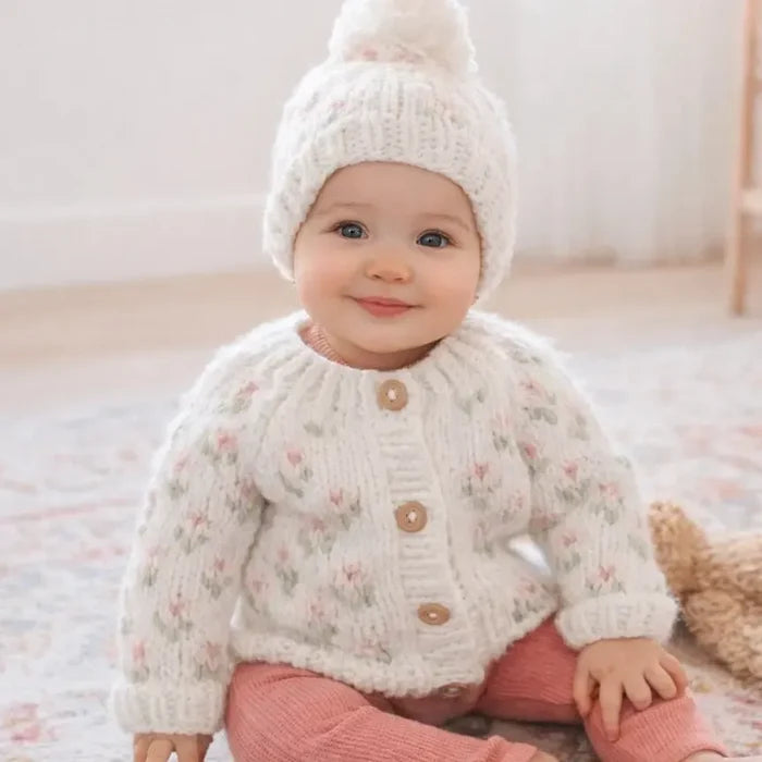 Baby wearing a knitted sweater and hat sitting on a soft surface.