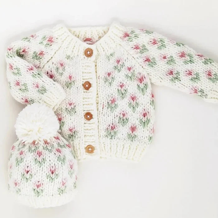 A cream knit baby cardigan and matching hat set with a pink and green floral pattern, displayed on a white background with a black banner at the top reading 'search ’bitty blooms’ to find the coordinating sweater'.