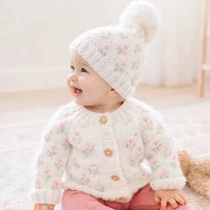 A baby sitting on a patterned rug wearing a cream knit cardigan and matching pom-pom hat with a small pink and green floral pattern. The baby is also wearing pink pants.