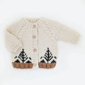 A cream-colored hand-knit baby cardigan featuring a cable knit pattern, three wooden buttons, and a Fair Isle style tree design in black and brown at the hem.