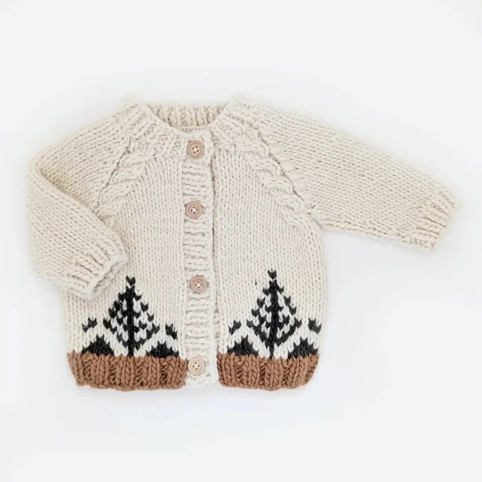 A cream-colored hand-knit baby cardigan featuring a cable knit pattern, three wooden buttons, and a Fair Isle style tree design in black and brown at the hem.