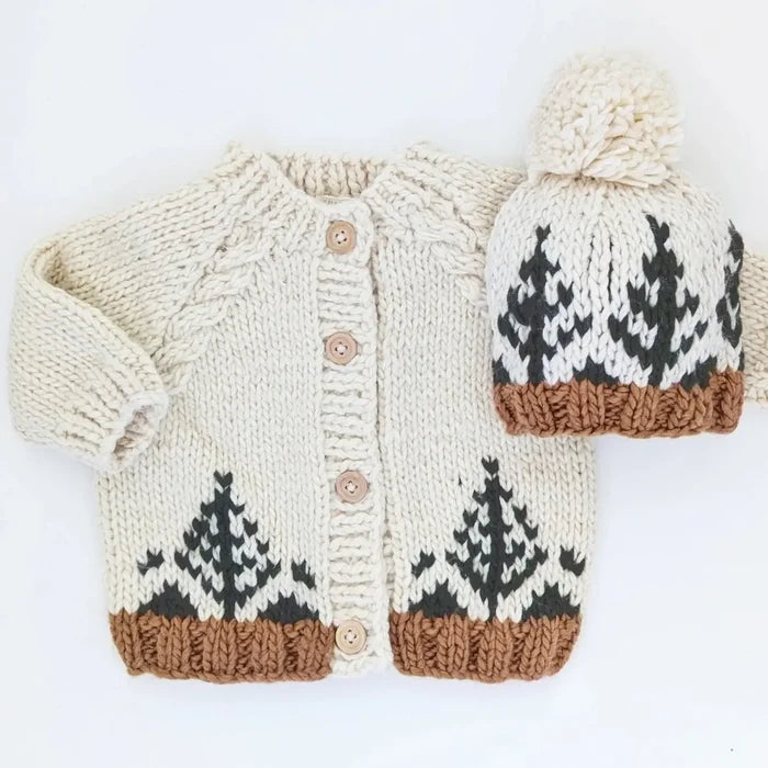 A cream knit baby cardigan and matching beanie hat with a forest tree pattern, displayed flat on a white background with a black banner at the top reading 'search 'forest' to find the coordinating hat'.