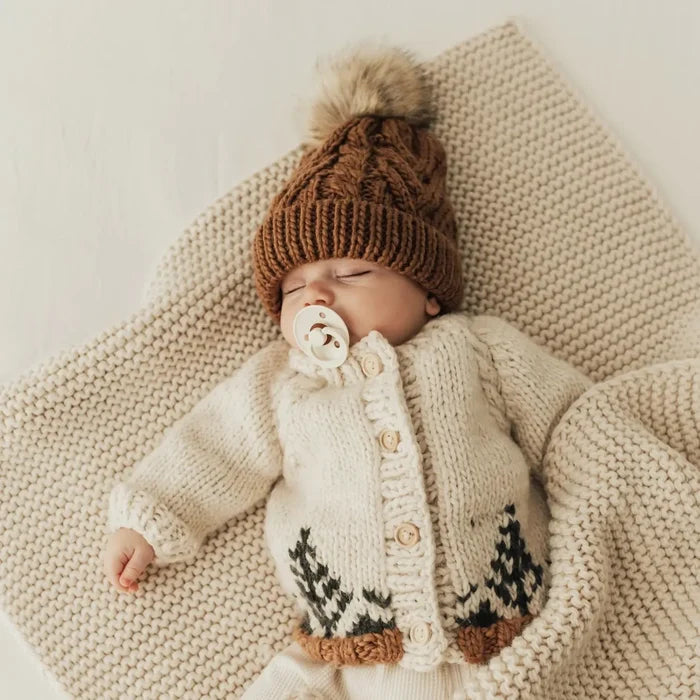 Newborn baby wearing a knitted brown hat and cream cardigan with deer pattern, lying on a textured blanket.
