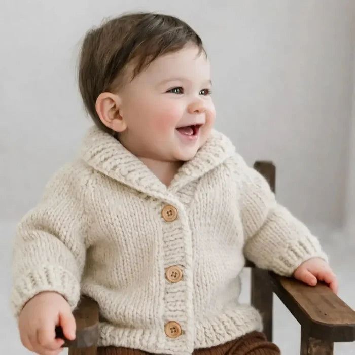 Baby wearing a beige knitted cardigan with brown buttons, sitting on a wooden stool against a plain background.