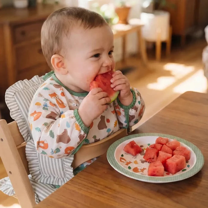 Baby eating watermelon in a high chair with a plate of watermelon slices on a wooden table.
