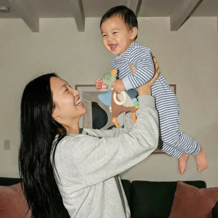 A woman in a light gray sweatshirt and matching pants holds a smiling baby up in a living room. The baby is wearing a blue and white striped pajama set.