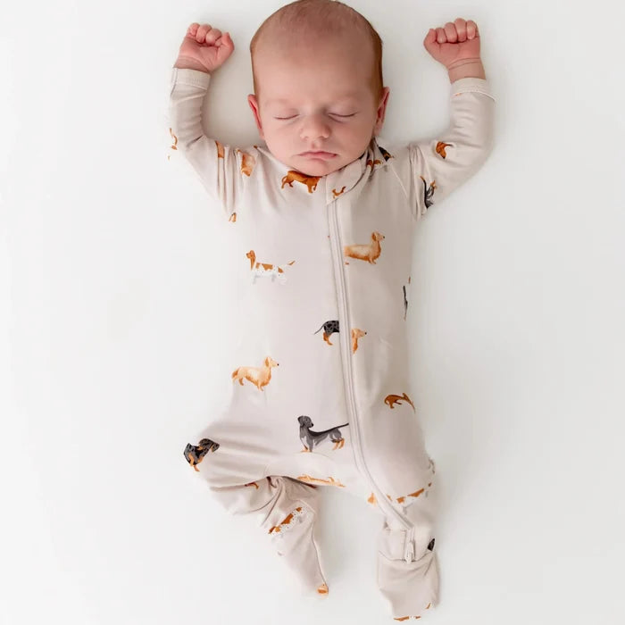 A sleeping newborn baby wearing a light beige footed sleeper with a print of small dachshund dogs, photographed from above on a clean white background.