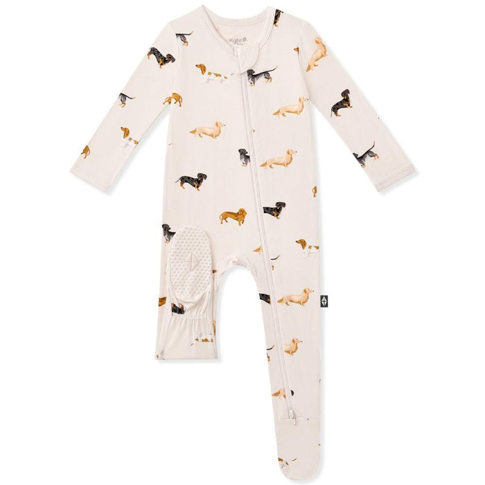 A baby footed sleeper in a warm cream color, featuring an all-over print of dachshunds in black, tan, and brown.