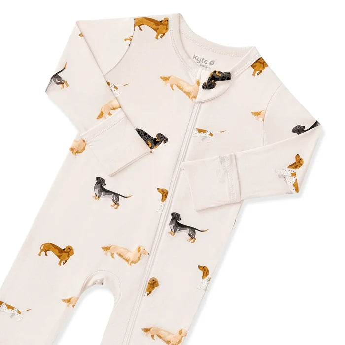 A long-sleeve baby footed sleeper in cream with an all-over print of dachshunds in tan, black, and cream, shown flat on a white background.