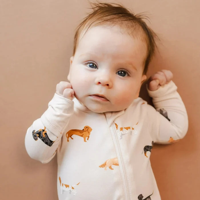 A baby wearing a white long-sleeve footless onesie with a print of dachshunds and other small dog illustrations, lying on a solid tan background.