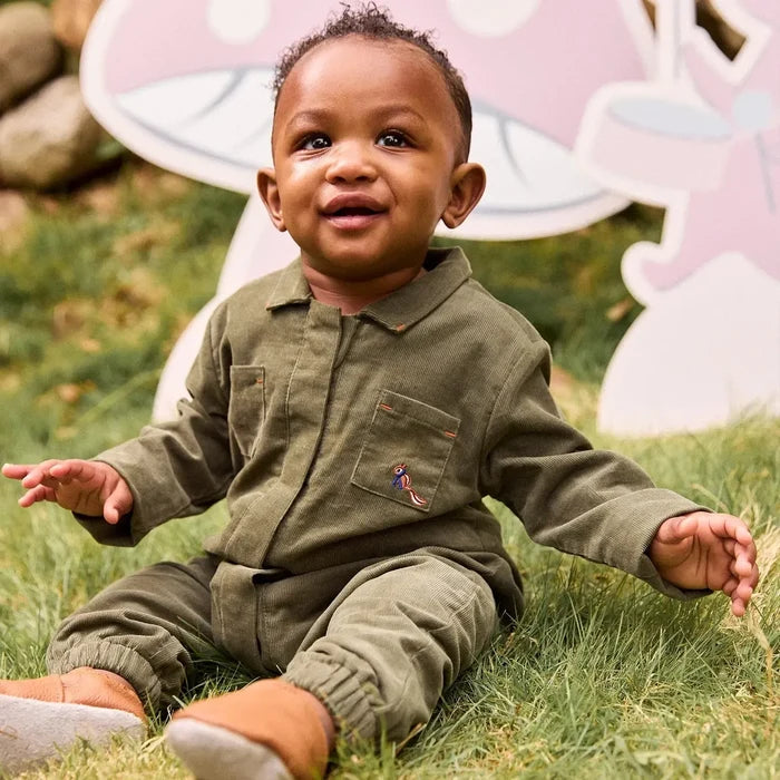 A smiling toddler sitting on green grass wearing an olive green corduroy one-piece jumpsuit with a small colorful embroidered squirrel on the pocket. A large pink and white cartoon decoration is visible in the background.