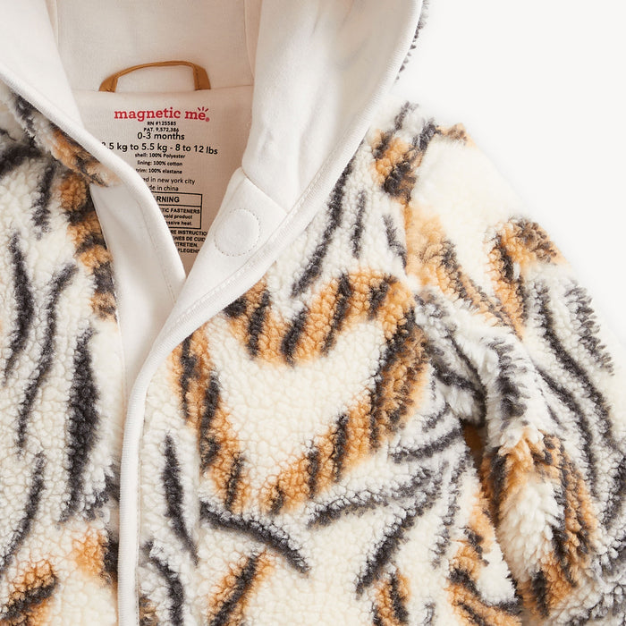 Close-up of a plush sherpa romper a tiger-stripe pattern in cream, tan, and dark gray/black. The inner collar shows the brand name 'magnetic me' and size information.