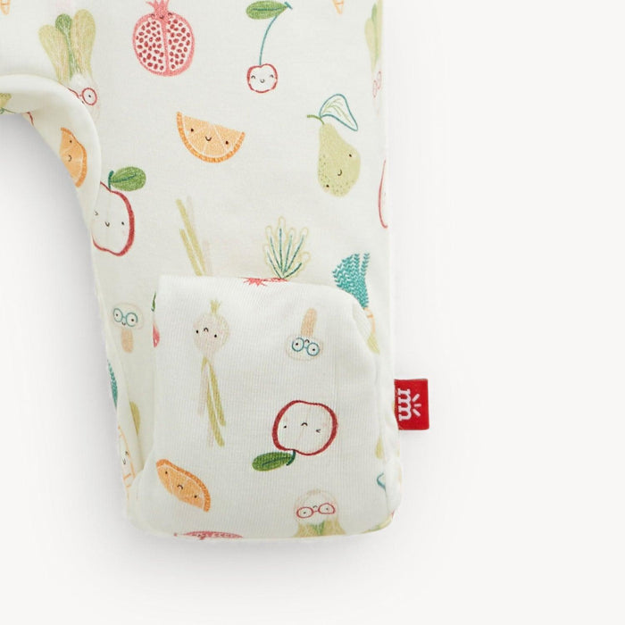 A close-up of a baby romper featuring a cute, colorful cartoon fruit and vegetable print on a warm off-white background. The fabric has a soft texture, and a small red fabric brand tag is visible on the right side.