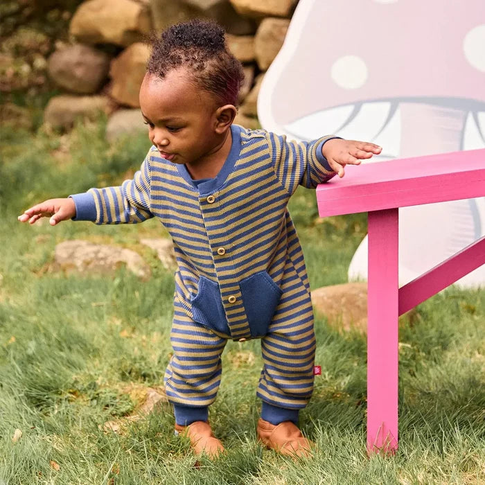 A toddler wearing a blue and olive yellow striped footless onesie stands on grass next to a pink table, holding onto a mushroom-shaped play structure.