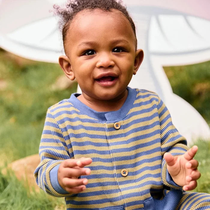 Image of a baby wearing a blue and olive striped romper, sitting outside in the grass