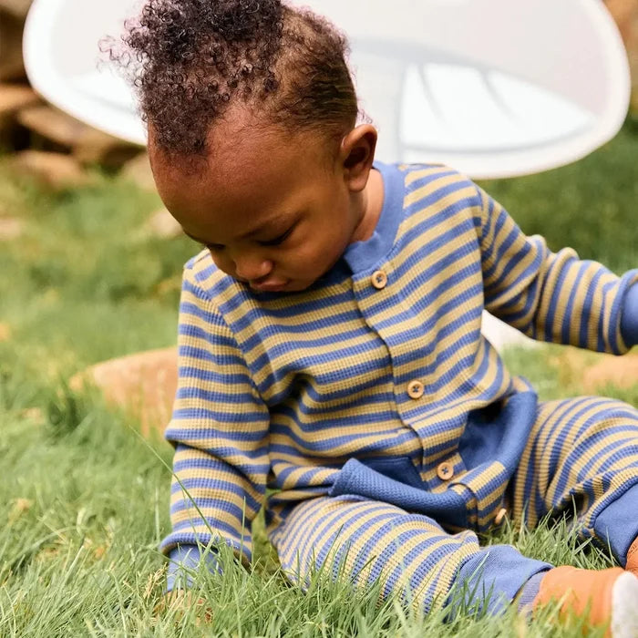 Image of a baby wearing a blue and olive striped romper, sitting outside in the grass