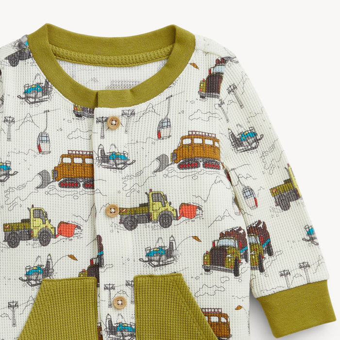 A close-up of a baby romper featuring an off-white waffle-knit body with a colorful winter mountain and ski lift print, olive green ribbed collar, cuffs, and pocket trims, and wooden buttons.