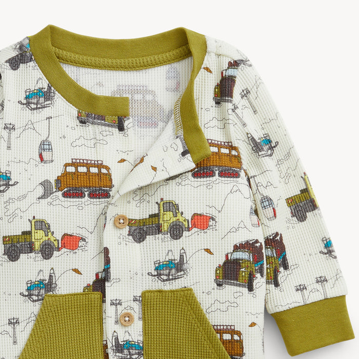 A close-up of a baby romper featuring an all-over construction truck print on a cream waffle-knit fabric, with olive green trim, collar, cuffs, and front pockets.